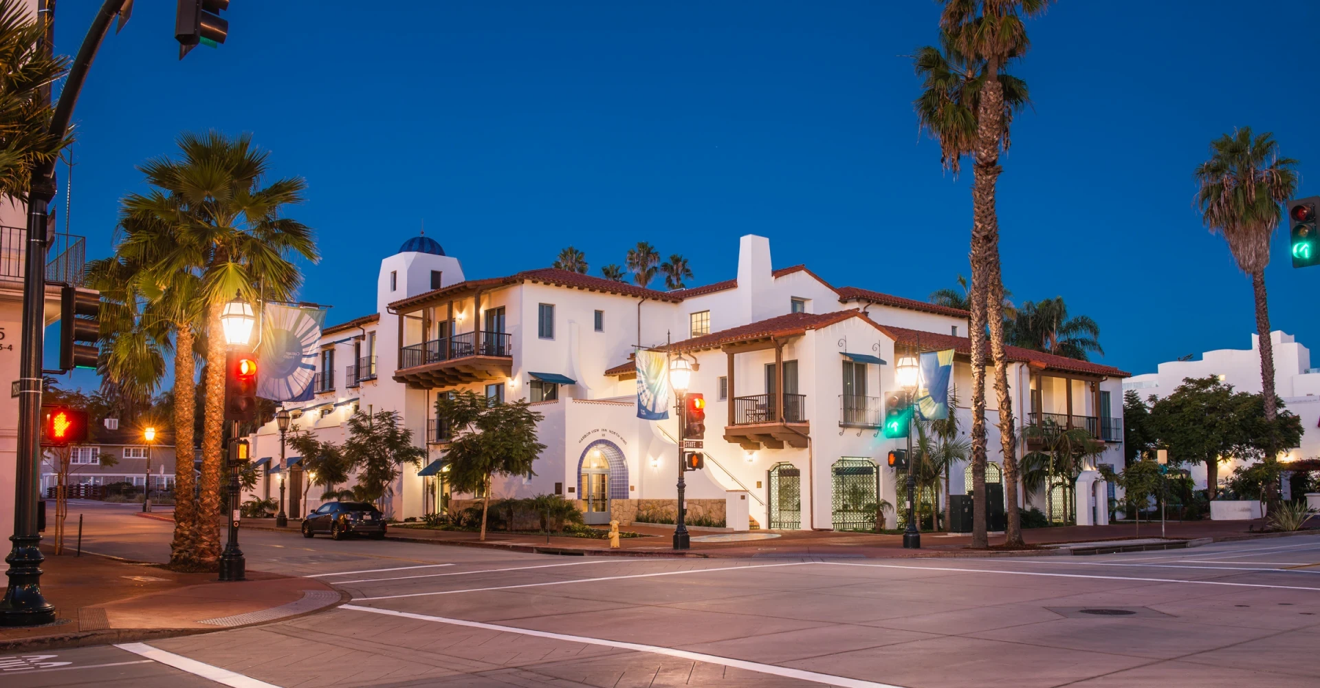 A white building with a red-tiled roof and palm trees in front of it at dusk.