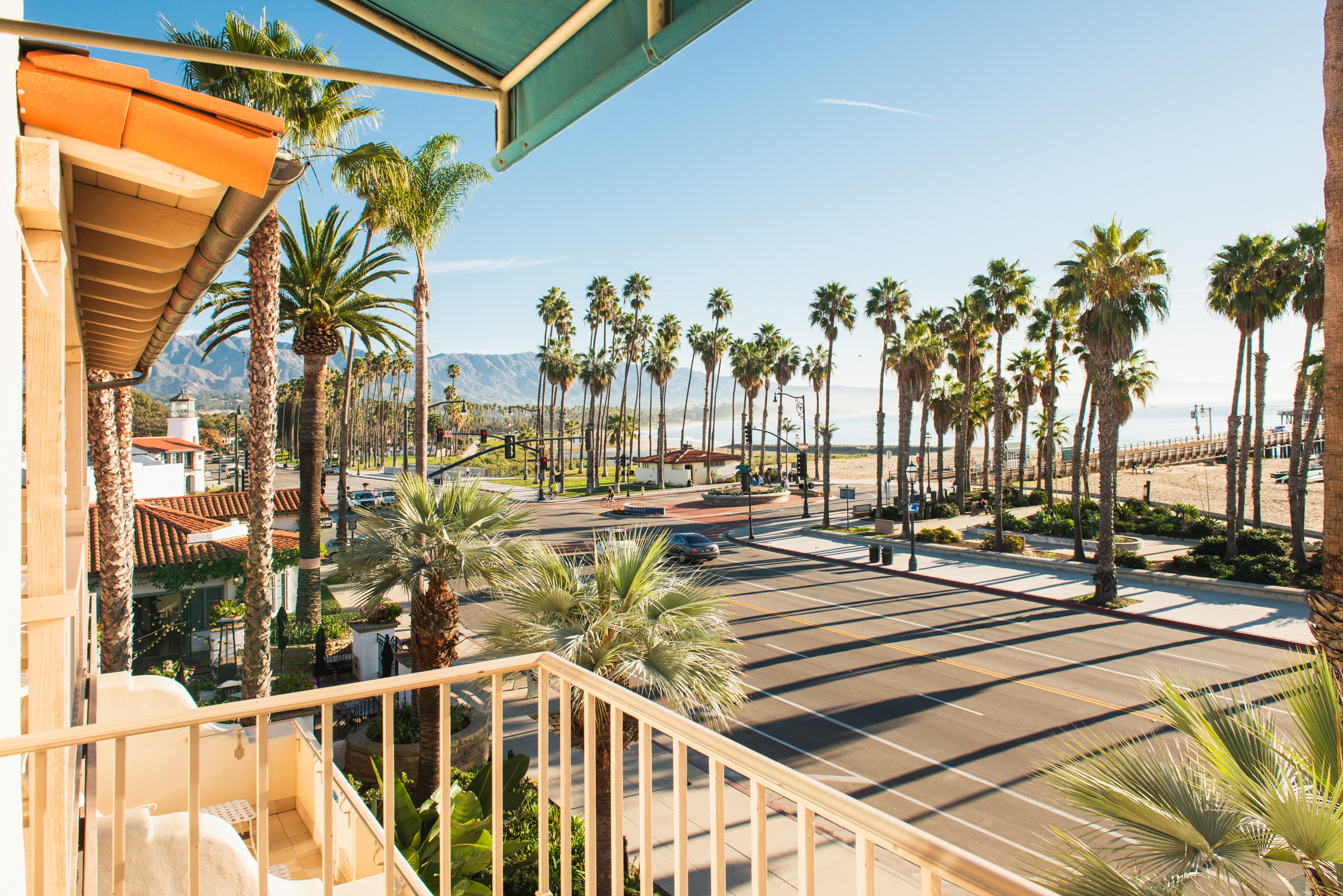 A view from a balcony of a palm-lined street leading to a beach with mountains in the distance.